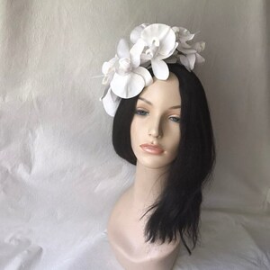 May include: White flower headband with three large white orchid flowers. The headband is made of a white material and is worn on a mannequin with black hair.