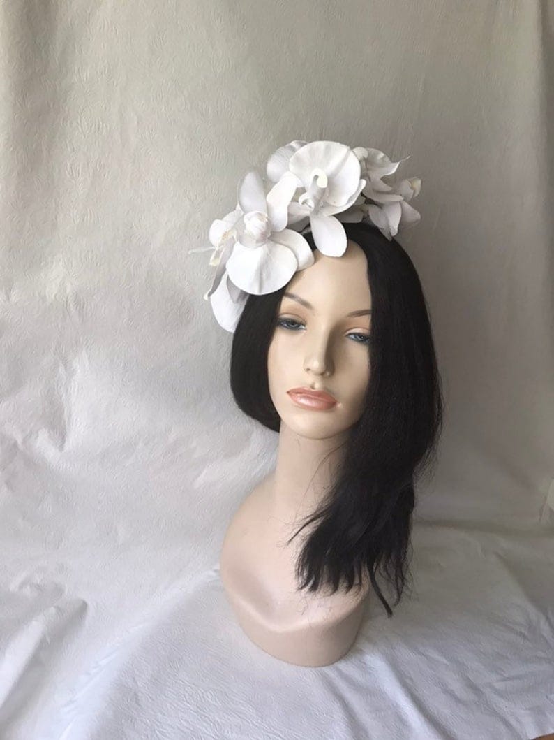 May include: White flower headband with three large white orchid flowers. The headband is made of a white material and is worn on a mannequin with black hair.