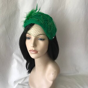 May include: A green felt hat with a beaded design and a small feather accent. The hat has a veil attached to it.