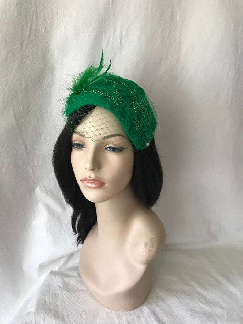 May include: A green felt hat with a beaded design and a small feather accent. The hat has a veil attached to it.