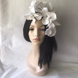 May include: A white flower headpiece with a cluster of white orchid blossoms. The headpiece is worn on a mannequin with long black hair.