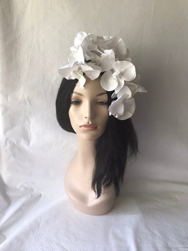 May include: A white flower headpiece with a cluster of white orchid blossoms. The headpiece is worn on a mannequin with long black hair.