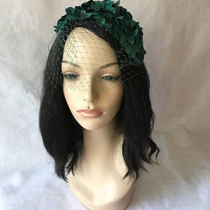 May include: A dark green floral headpiece with a matching veil. The headpiece is adorned with fabric flowers and small embellishments. The veil is a fine mesh, adding a vintage touch to the design. The headpiece is displayed on a mannequin head.