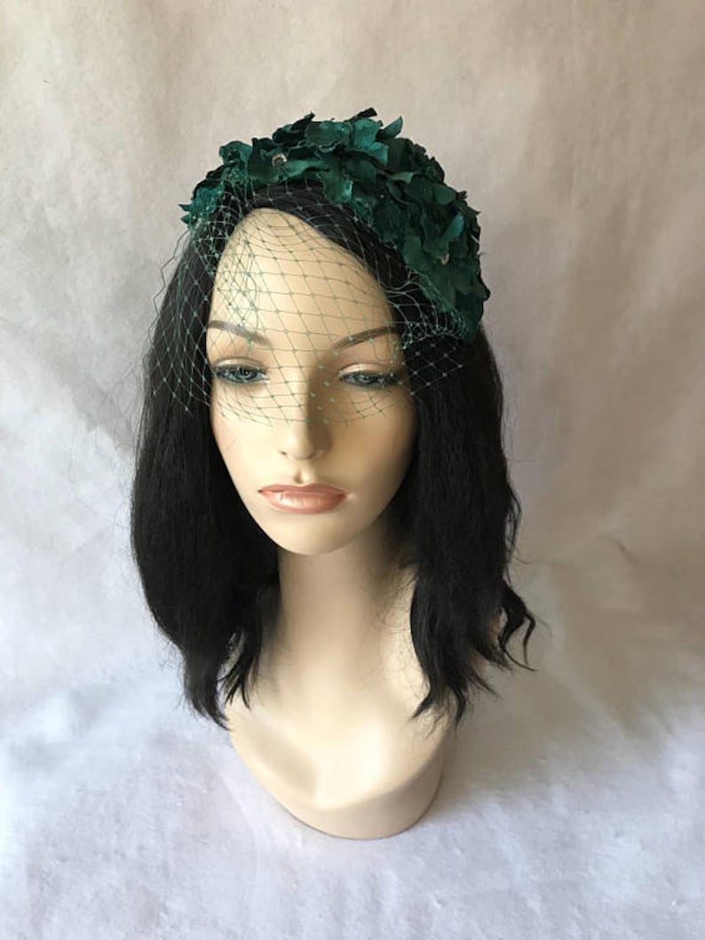 May include: A dark green floral headpiece with a matching veil. The headpiece is adorned with fabric flowers and small embellishments. The veil is a fine mesh, adding a vintage touch to the design. The headpiece is displayed on a mannequin head.