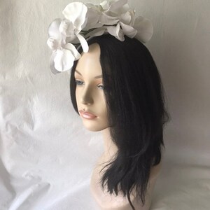 May include: White flower headband with a large white orchid design. The headband is made of a white material and is worn on a mannequin with black hair.