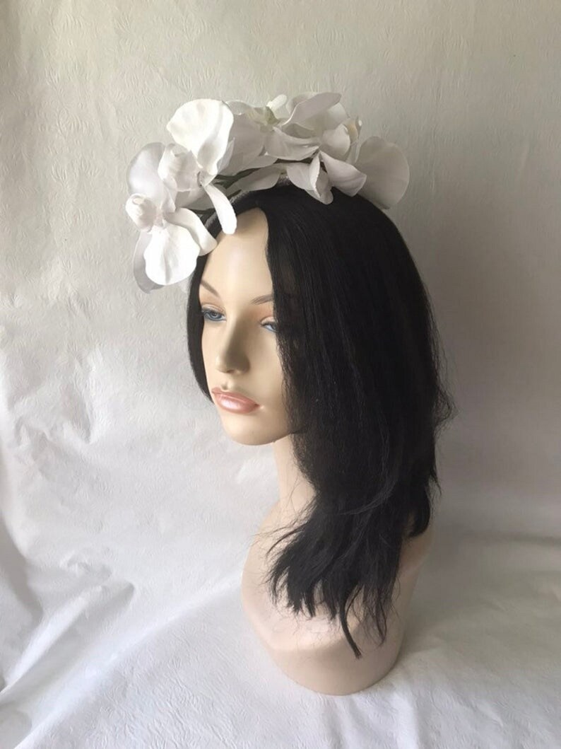 May include: White flower headband with a large white orchid design. The headband is made of a white material and is worn on a mannequin with black hair.