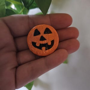 May include: A small, round, orange-painted object shaped like a jack-o'-lantern. It features a glossy finish with black painted details, including two triangular eyes, a small triangular nose, and a wide, toothy grin carved into the surface.