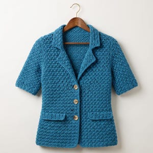 Op de afbeelding: Een blazer met korte mouwen gemaakt van blauw garen met een gehaakt patroon met textuur. Het kledingstuk heeft een reverskraag, drie lichtgekleurde ronde knopen aan de voorzijde en twee rechthoekige klepzakken op de taillehoogte.