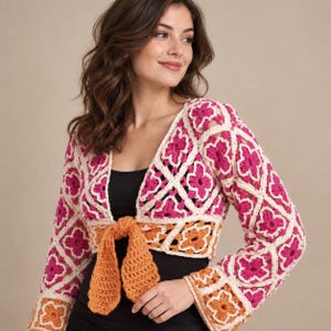 Puede incluir: Un bolero de ganchillo de manga larga con un motivo floral repetitivo en magenta vibrante y crema, con un lazo de ganchillo naranja sólido en la cintura. La prenda tiene una silueta corta y se lleva sobre una camiseta negra sencilla.
