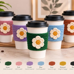 Pode incluir: Cinco copos de café descartáveis brancos com tampas pretas, cada um com uma manga de crochê em rosa, roxo, verde escuro, azul claro ou ferrugem. Cada manga é decorada com uma aplicação central de flor de crochê branca e amarela.