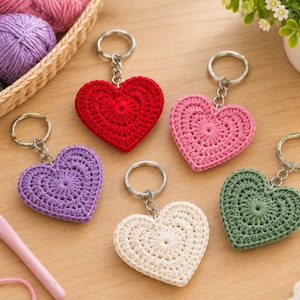 May include: Five heart-shaped keychains made of crocheted yarn in red, pink, purple, cream, and green. Each keychain features a silver-toned metal ring and chain attached to the top. A pink crochet hook lies nearby on the surface.