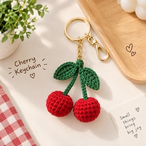 May include: A crochet cherry keychain featuring two round red yarn spheres attached to green stems and leaves. The keychain includes a gold-toned metal ring and a lobster clasp. The text "Cherry Keychain" and "Small things bring big joy" are visible.
