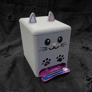 May include: A light grey rectangular dispenser featuring a cat face design with two pointed ears, black eyes, a pink nose, whiskers, and two black paw print cutouts at the bottom. A purple and blue rectangular packet is partially inserted at the base.