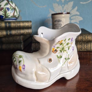 May include: A white ceramic shoe-shaped planter featuring hand-painted floral motifs in purple, yellow, and green. The glossy finish highlights the moulded details of the boot, including eyelets and a curved opening, creating a decorative vessel.