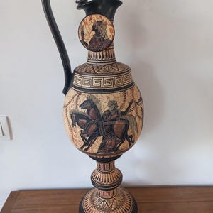 May include: A tall ceramic pitcher with a black handle and base, featuring cream-colored surfaces decorated with black and reddish-brown illustrations. The design includes a chariot with two horses, geometric Greek key patterns, and a circular portrait.