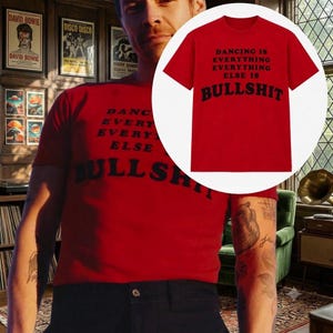May include: A bright red short-sleeved t-shirt featuring black text printed on the front that reads: DANCING IS EVERYTHING EVERYTHING ELSE IS BULLSHIT. The shirt has a crew neckline and a standard fit, shown both on a person and in a circular inset.