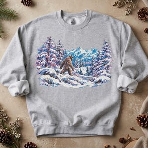 May include: A light gray crewneck sweatshirt featuring a graphic print of a brown Bigfoot creature walking through a snowy mountain landscape with evergreen trees, blue peaks, and white snow-covered ground under a clear sky.