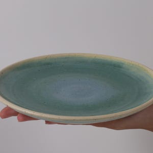 May include: A round ceramic plate featuring a gradient glaze that transitions from a deep blue center to a soft teal rim. The surface has a speckled texture throughout, and the outer edge is finished with a light cream-colored border.