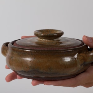 May include: A round, brown ceramic casserole dish with a matching lid and two side handles. The glaze features a mottled, earthy texture with darker brown and tan tones, showcasing a smooth, glossy finish on the exterior and lid surface.