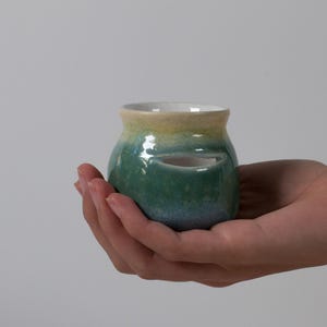 May include: A small, rounded ceramic pot with a glossy finish that transitions from a pale yellow at the rim to a deep teal and blue at the base. The vessel features a distinct horizontal slit opening on its side and a white interior.