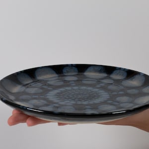 May include: A round ceramic plate featuring a glossy dark navy blue and black glaze. The surface is decorated with a repeating pattern of light blue and white circular spots that radiate from the center toward the outer rim.