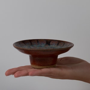 May include: A small, round ceramic bowl with a pedestal base. The exterior features a mottled, warm reddish-brown glaze, while the interior displays a deep blue and brown reactive glaze pattern with a smooth, glossy finish.