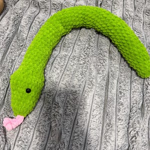 May include: A long, curved, lime green plush snake toy made of soft, chunky yarn. The snake features a textured, crocheted surface, a small black bead eye on the side of its head, and a bifurcated pink tongue protruding from its mouth.