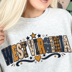 Peut inclure: Un sweat-shirt gris clair à col rond arborant le texte WEST VIRGINIA dans une police audacieuse à motifs multiples. Les lettres intègrent des designs à carreaux et géométriques bleu marine, jaune moutarde et beige, agrémentés d'étoiles.