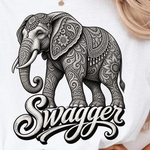 May include: A white t-shirt featuring a black and white graphic illustration of an elephant with intricate mandala-style patterns on its body. Below the elephant, the word "Swagger" is printed in a bold, cursive, black script font.