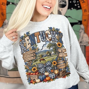 Peut inclure: Un sweat-shirt gris clair à col rond avec une impression graphique portant le mot KENTUCKY en lettres à carreaux bleus et blancs. Le motif comprend un paysage avec un cheval, un tonneau, des citrouilles, des pommes, une lanterne et un chapeau de cowboy.