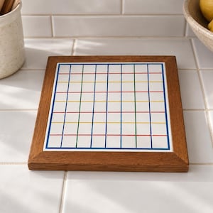 May include: A square wooden trivet featuring a white ceramic tile insert. The tile is printed with a grid pattern composed of thin blue, green, and red lines, creating a series of small squares across the surface of the kitchen accessory.