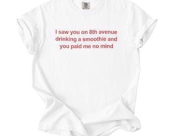 May include: A white short-sleeve t-shirt featuring red text centered on the chest that reads: I saw you on 8th avenue drinking a smoothie and you paid me no mind. The garment has a crew neckline and a standard fit with rolled sleeve cuffs.