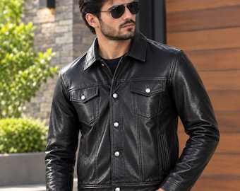 May include: A black leather jacket featuring a classic collar, long sleeves, and a front button-down closure. The garment includes two chest pockets with buttoned flaps and vertical seam detailing, providing a structured fit.