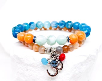 Puede incluir: Dos pulseras superpuestas con cuentas azules, verdes y naranjas. La pulsera superior tiene un dije con un símbolo Om y cuentas azules, rojas y negras.