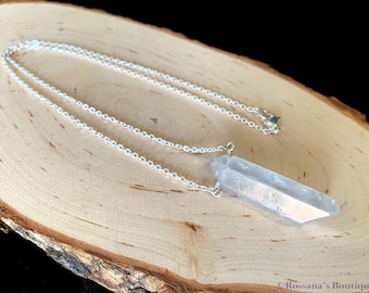 Puede incluir: Un collar de cadena de plata con un colgante de cristal de cuarzo transparente. El cristal está facetado y tiene un ligero brillo iridiscente.