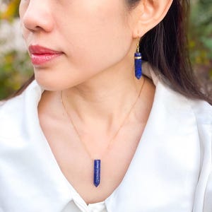 May include: A gold chain necklace with a long, pointed, dark blue gemstone pendant. Matching earrings with a similar gemstone dangle from the wearer's ear.