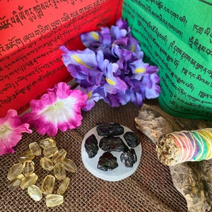 Puede incluir: Una colecci&oacute;n de art&iacute;culos espirituales que incluyen un palo de salvia blanca, un peque&ntilde;o cuenco de cristales negros, una pila de piedras de citrino amarillo y dos banderas de oraci&oacute;n con texto tibetano. Las banderas de oraci&oacute;n son rojas y verdes con texto blanco.