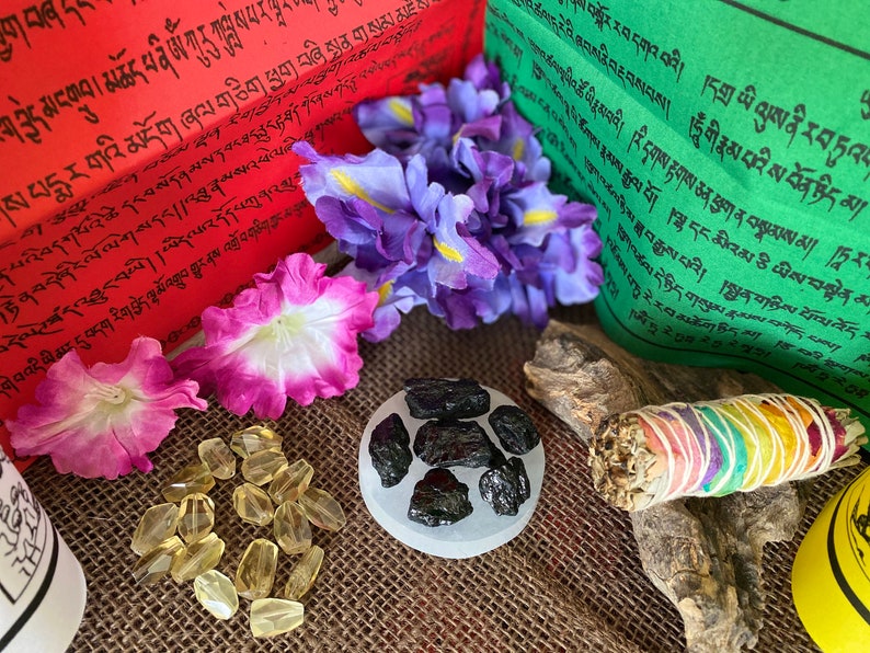 Puede incluir: Una colecci&oacute;n de art&iacute;culos espirituales que incluyen un palo de salvia blanca, un peque&ntilde;o cuenco de cristales negros, una pila de piedras de citrino amarillo y dos banderas de oraci&oacute;n con texto tibetano. Las banderas de oraci&oacute;n son rojas y verdes con texto blanco.