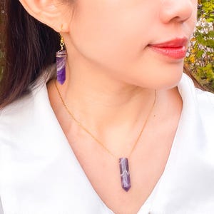 May include: A gold chain necklace with a purple amethyst pendant. The pendant is a long, pointed crystal. The necklace is worn by a person with a matching pair of amethyst earrings.