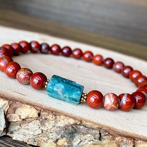Puede incluir: Un brazalete de cuentas de jaspe rojo con una cuenta central de apatita azul turquesa. El brazalete est&aacute; sobre una superficie de madera.