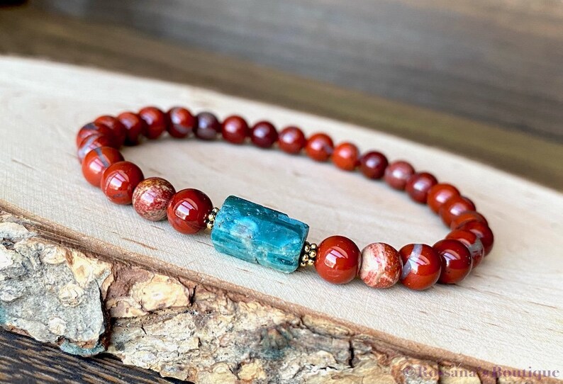 Puede incluir: Un brazalete de cuentas de jaspe rojo con una cuenta central de apatita azul turquesa. El brazalete est&aacute; sobre una superficie de madera.