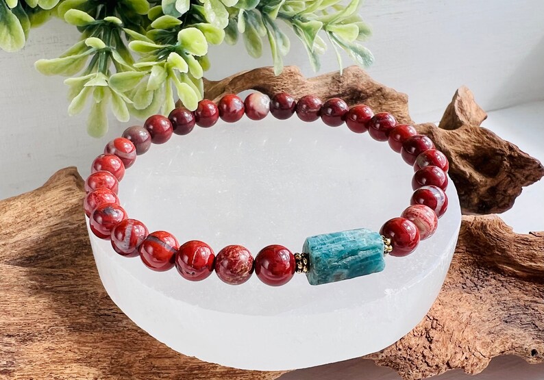 Puede incluir: Pulsera de cuentas de jaspe rojo con un acento de piedra preciosa azul turquesa. La pulsera est&aacute; sobre una placa de carga de selenita blanca.