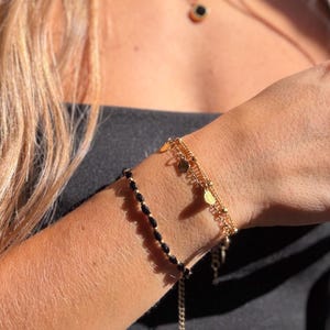 May include: A layered bracelet set consisting of one thin black beaded cord bracelet and one gold-toned chain bracelet featuring small round charms and beads. The gold chain has a delicate link structure with evenly spaced metallic accents.