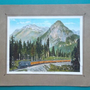 May include: Vintage print of a train traveling through a mountain landscape. The image features a train with orange passenger cars, set against a backdrop of green trees, a blue sky, and a large mountain. The print is framed in a brown border.