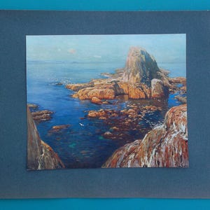 May include: A framed landscape painting depicting a coastal scene. The artwork features a rocky shoreline with a large rock formation in the center, surrounded by blue water and a light blue sky. The painting is mounted on a dark blue mat.