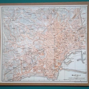 Puede incluir: Un mapa antiguo de Nápoles, Italia, enmarcado en un borde marrón. El mapa presenta planos de calles detallados, características geográficas y la costa. El mapa está en tonos marrones y blancos, con el nombre de la ciudad "Napoli" en la parte inferior.