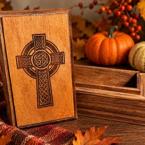 May include: A rectangular wooden box with a removable lid featuring a dark brown laser-engraved Celtic cross design. The box is constructed from light and dark wood tones, showcasing a natural grain pattern on the lid and the open storage compartment.
