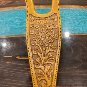 May include: A tan-colored boot jack featuring a U-shaped top and a tapered base. The surface is decorated with an embossed floral pattern and the word "acme" stamped near the top. The edges are detailed with a series of small, evenly spaced indentations.