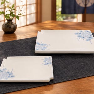 May include: Two rectangular white ceramic plates featuring a delicate blue floral pattern painted in opposite corners. Each plate has a smooth, glossy finish and a notched edge detail on one side, presenting a clean and functional design for serving.