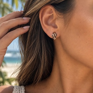 May include: A small stud earring shaped like a spiral seashell with a cream-colored base and dark brown checkered or spotted markings. The earring is secured to the earlobe with a metallic post, featuring a detailed, textured surface.